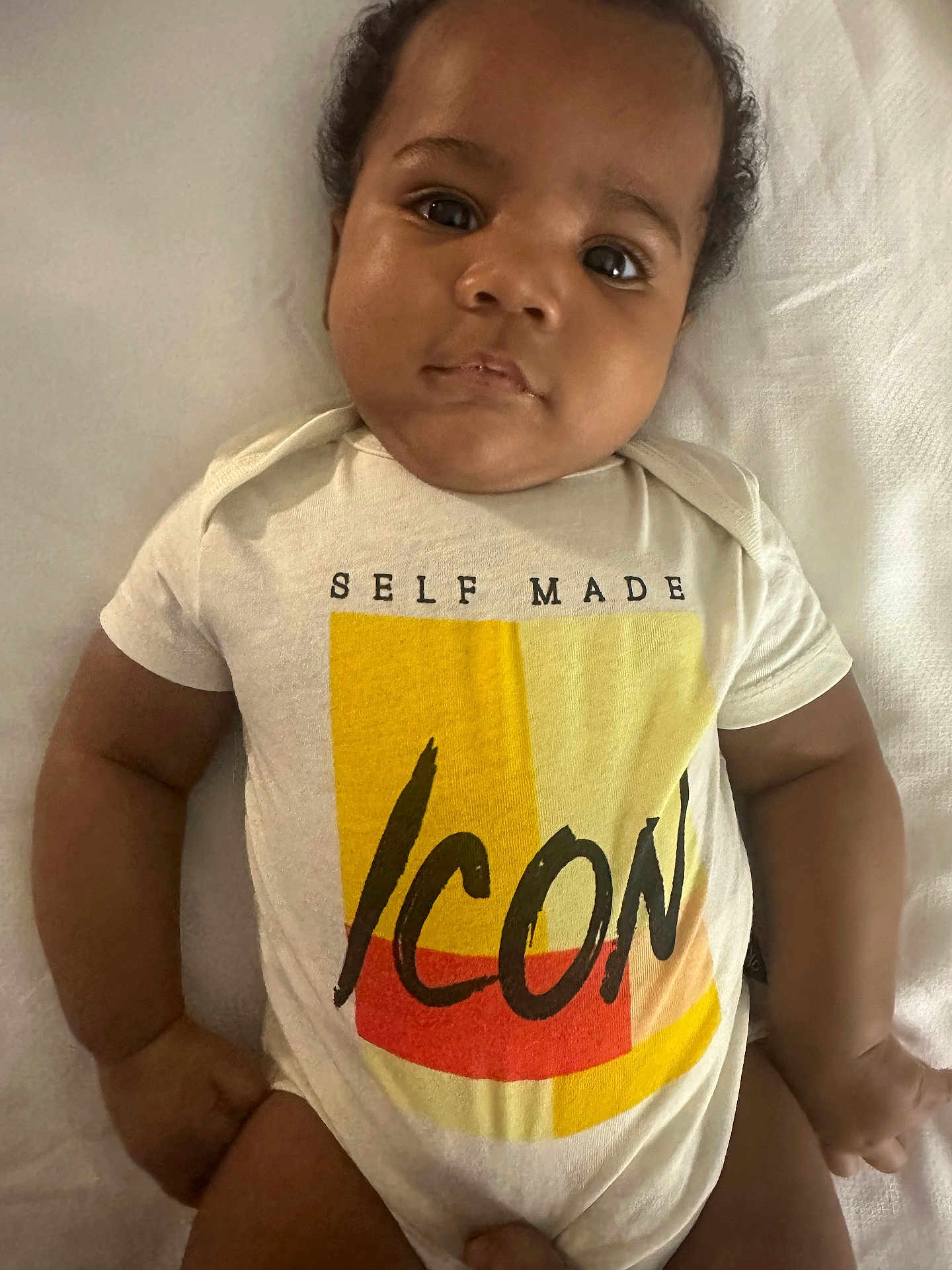 Jayceon is registered to the contest to win money with this photo: baby, infant, child, onesie, clothing, text_on_clothing, face, arms, legs, skin, lying_down, blanket, indoors, portrait, cute, young, expression, closeup, person, toddler