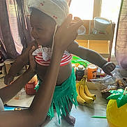 Akiyra is registered to the contest to win money with this photo: toddler, child, baby, hat, kitchen, bananas, snacks, pineapple_cup, table, curtains, person, arms, clothing, green_skirt, red_stripes, hands, indoor, food, fruit, standing