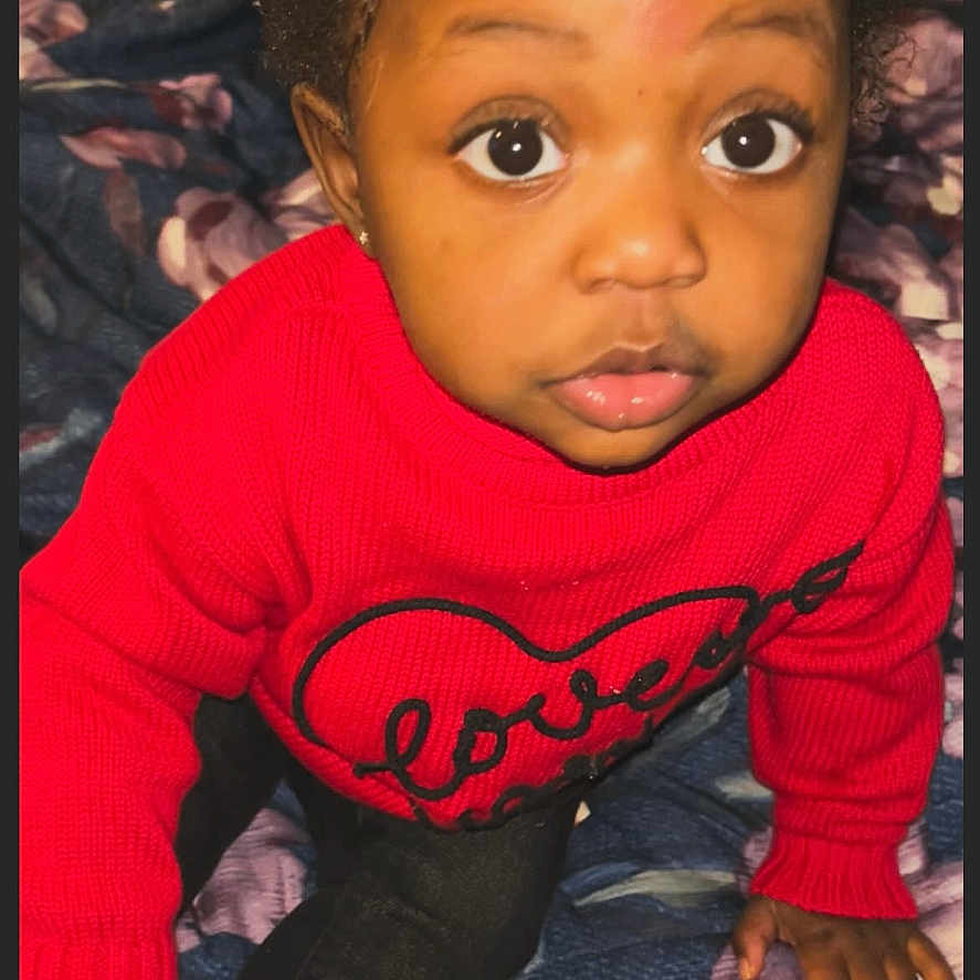 Kinzey is registered to the contest to win money with this photo: big_eyes, child, closeup, clothing, cozy, curly_hair, cute, expression, floral_blanket, hair_accessories, hand, heart_design, indoor, person, portrait, red_sweater, sitting, toddler, warm_clothes, young_child