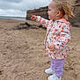 toddler, child, beach, sand, jacket, butterfly_pattern, purple_pants, white_shoes, pointing, side_profile, outdoor, windy, hair, young_child, casual_clothing, daytime, cloudy_sky, person, footwear, fun