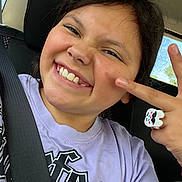 Liliana joined the competition — help win amazing prizes! child, smiling, portrait, selfie, car, car_seat, seat_belt, indoors, vehicle, happy, teeth, face, dark_hair, purple_shirt, hand_gesture, ring, bracelet, window, daylight, playful
