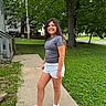 Liliana is registered to the contest to win money with this photo: child, girl, smiling, portrait, outdoor, sidewalk, suburban, neighborhood, house, trees, grass, summer, casual_clothing, gray_shirt, white_shorts, sneakers, long_hair, standing, mailbox, daylight