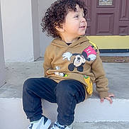 Anders is registered to the contest to win money with this photo: toddler, child, curly_hair, hoodie, mickey_mouse, sneakers, puma, pants, sitting, concrete, steps, outdoor, door, smiling, casual_clothing, side_view, young_child, person, daylight, happy