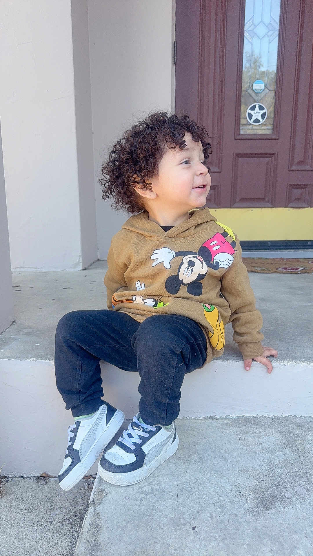 Anders is registered to the contest to win money with this photo: toddler, child, curly_hair, hoodie, mickey_mouse, sneakers, puma, pants, sitting, concrete, steps, outdoor, door, smiling, casual_clothing, side_view, young_child, person, daylight, happy