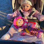 Niamh joined the competition — help win amazing prizes! child, toddler, stroller, pink_jacket, hat, doll, toy, blanket, outdoor, sunlight, shadow, person, baby, seatbelt, cold_weather, cute, portrait, playful, happy, face
