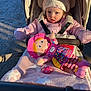 child, toddler, stroller, pink_jacket, hat, doll, toy, blanket, outdoor, sunlight, shadow, person, baby, seatbelt, cold_weather, cute, portrait, playful, happy, face