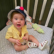 Leah is registered to the contest to win money with this photo: baby, hat, flowers, crib, pacifier, toy, smiling, happy, child, footwear, white_shoes, yellow_shirt, indoor, bedding, pattern, moon, plush_toy, person, cute, infant