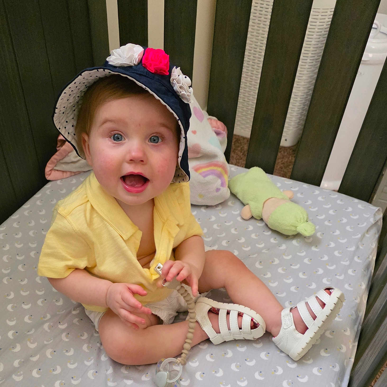 Leah is registered to the contest to win money with this photo: baby, bedding, child, crib, cute, flowers, footwear, happy, hat, indoor, infant, moon, pacifier, pattern, person, plush_toy, smiling, toy, white_shoes, yellow_shirt