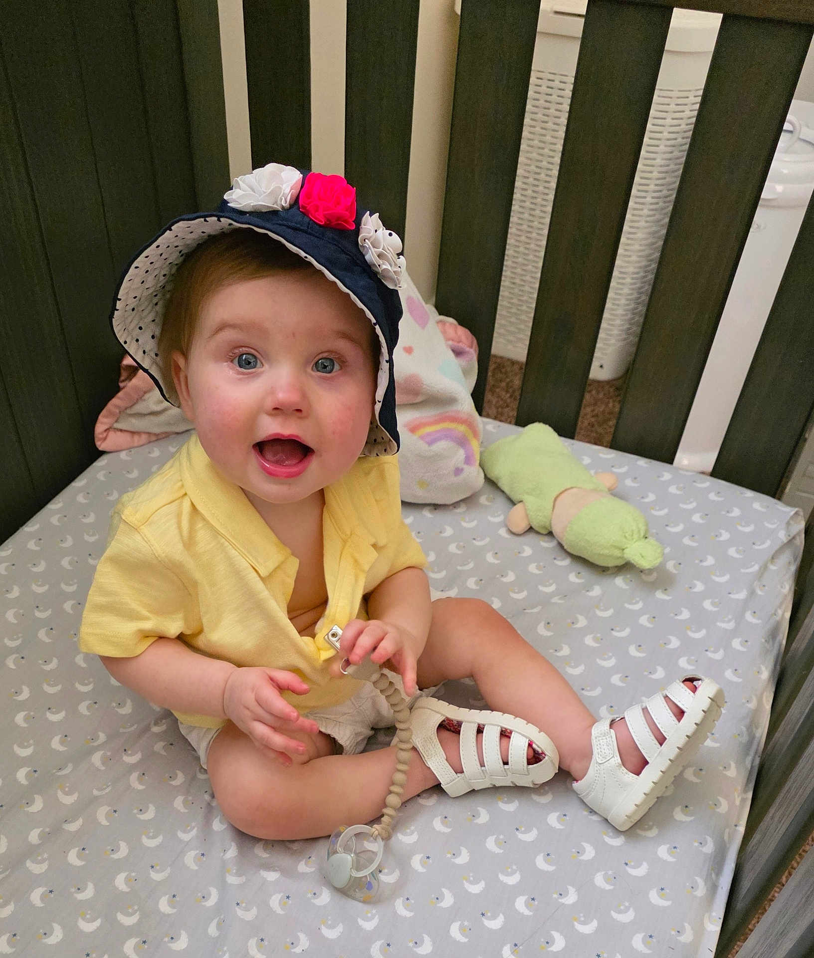Leah is registered to the contest to win money with this photo: baby, hat, flowers, crib, pacifier, toy, smiling, happy, child, footwear, white_shoes, yellow_shirt, indoor, bedding, pattern, moon, plush_toy, person, cute, infant