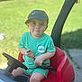 baby, backyard, cap, child, cute, daylight, grass, green_clothing, hat, outdoor, person, playful, red_toy, seatbelt, sitting, smile, steering_wheel, summer, toddler, toy_car