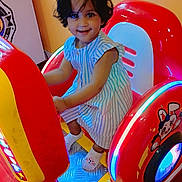 Misha participe au concours pour gagner de l'argent avec cette photo : child, toddler, arcade_machine, ride, smile, striped_dress, short_hair, socks, indoor, colorful, red, yellow, blue_lights, playful, fun, entertainment, cute, person, happy, toy