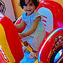 child, toddler, arcade_machine, ride, smile, striped_dress, short_hair, socks, indoor, colorful, red, yellow, blue_lights, playful, fun, entertainment, cute, person, happy, toy