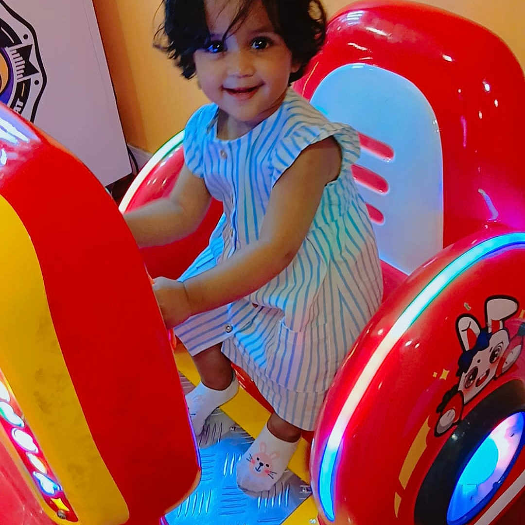 Misha participe au concours pour gagner de l'argent avec cette photo : arcade_machine, blue_lights, child, colorful, cute, entertainment, fun, happy, indoor, person, playful, red, ride, short_hair, smile, socks, striped_dress, toddler, toy, yellow