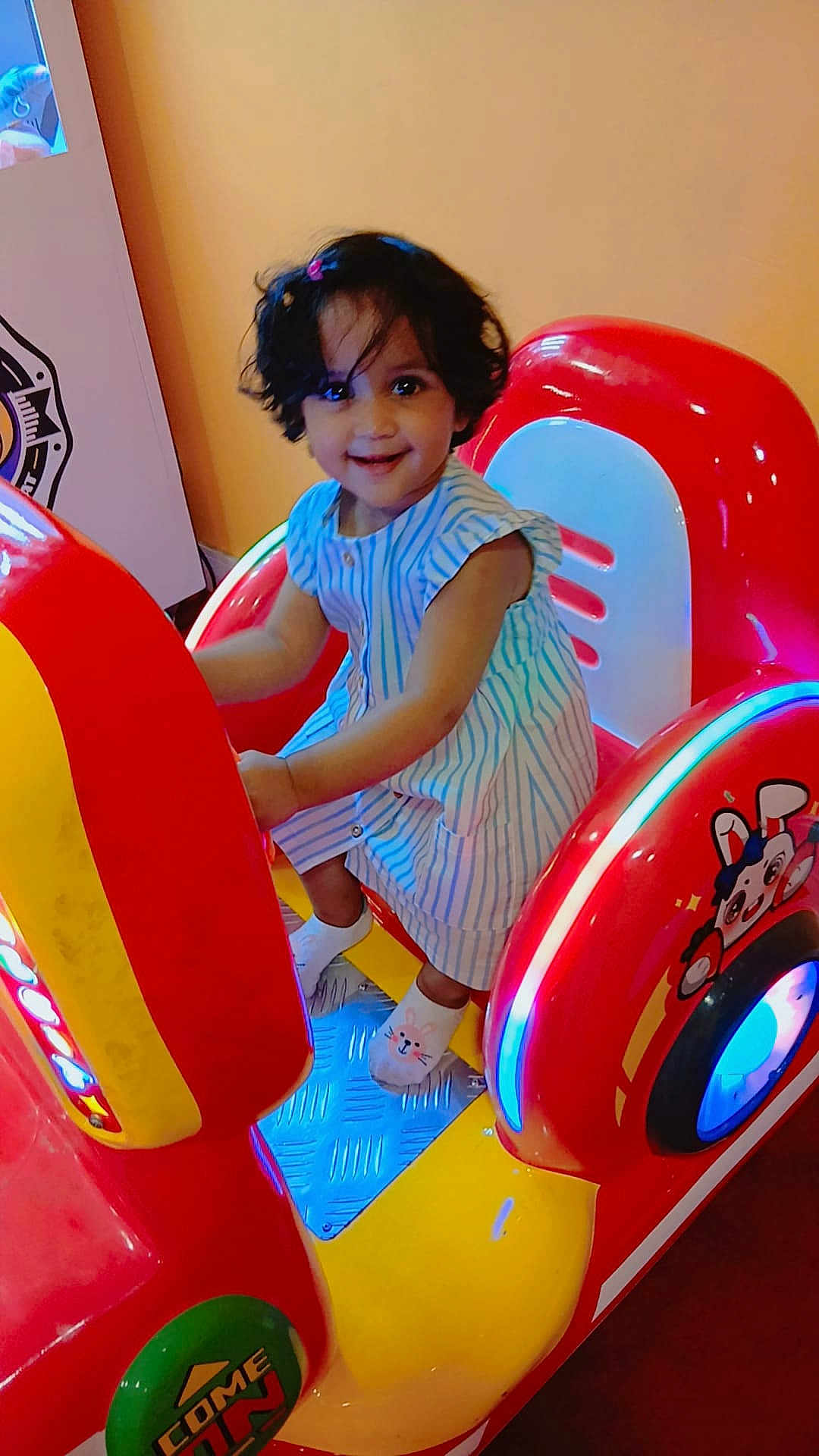 Misha participe au concours pour gagner de l'argent avec cette photo : child, toddler, arcade_machine, ride, smile, striped_dress, short_hair, socks, indoor, colorful, red, yellow, blue_lights, playful, fun, entertainment, cute, person, happy, toy
