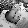 baby, infant, wicker_basket, plush_toy, stuffed_animal, pacifier, blanket, pillow, closeup, portrait, smiling, eyes, lying_down, nursery, soft_texture, black_and_white, monochrome, crib, head, cozy