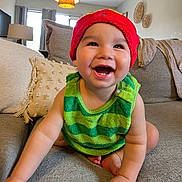 Josiah is registered to the contest to win money with this photo: baby, child, smiling, crawling, red_hat, green_top, couch, pillow, living_room, indoor, happy, cute, infant, face, skin, arm, foot, lamp, window, blanket