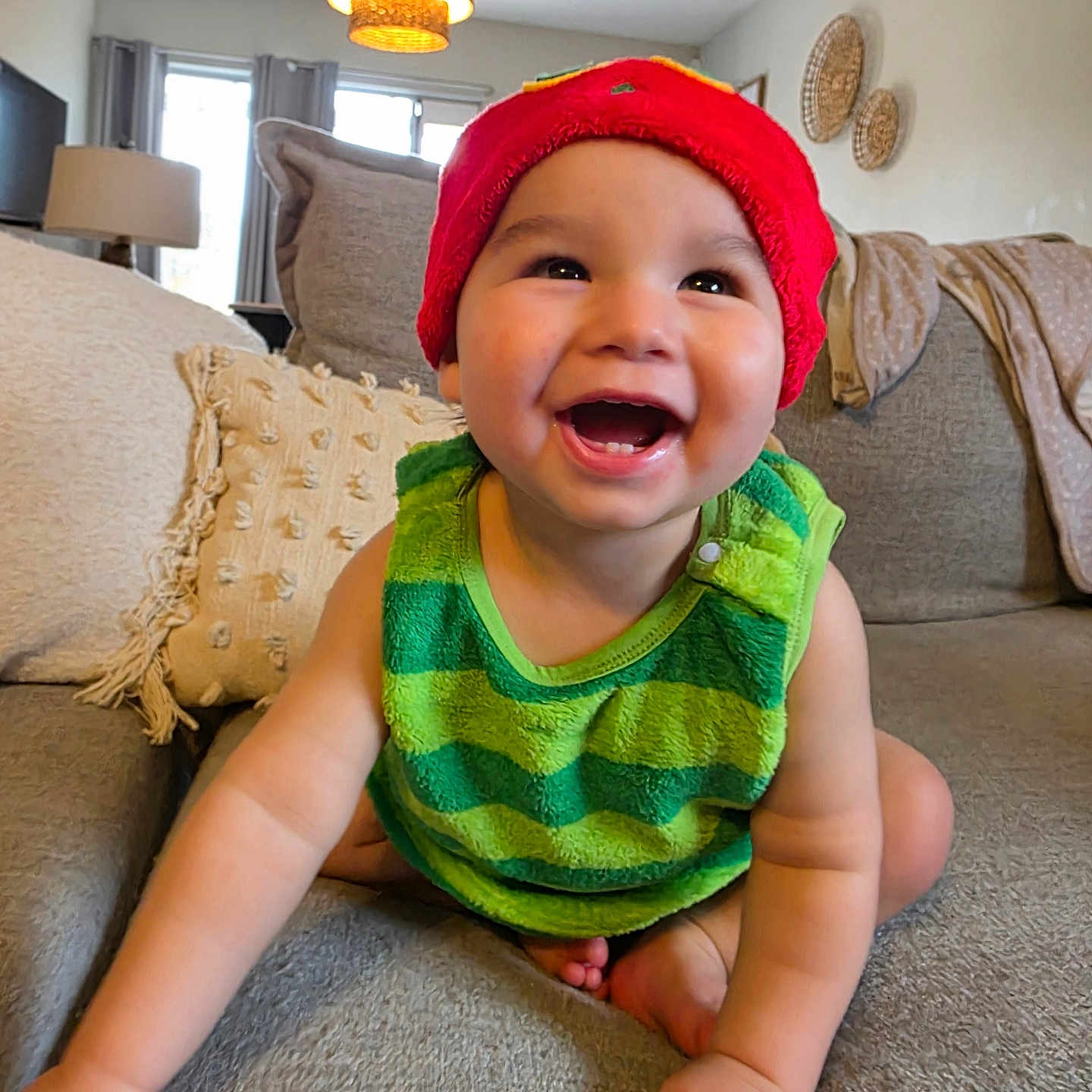 Josiah is registered to the contest to win money with this photo: baby, child, smiling, crawling, red_hat, green_top, couch, pillow, living_room, indoor, happy, cute, infant, face, skin, arm, foot, lamp, window, blanket