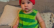 Josiah is registered to the contest to win money with this photo: toddler, child, hat, red_hat, green_outfit, striped_clothing, couch, living_room, pillow, lamp, indoor, person, baby, seated, cute, curious, furniture, home, casual, portrait