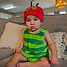 toddler, child, hat, red_hat, green_outfit, striped_clothing, couch, living_room, pillow, lamp, indoor, person, baby, seated, cute, curious, furniture, home, casual, portrait