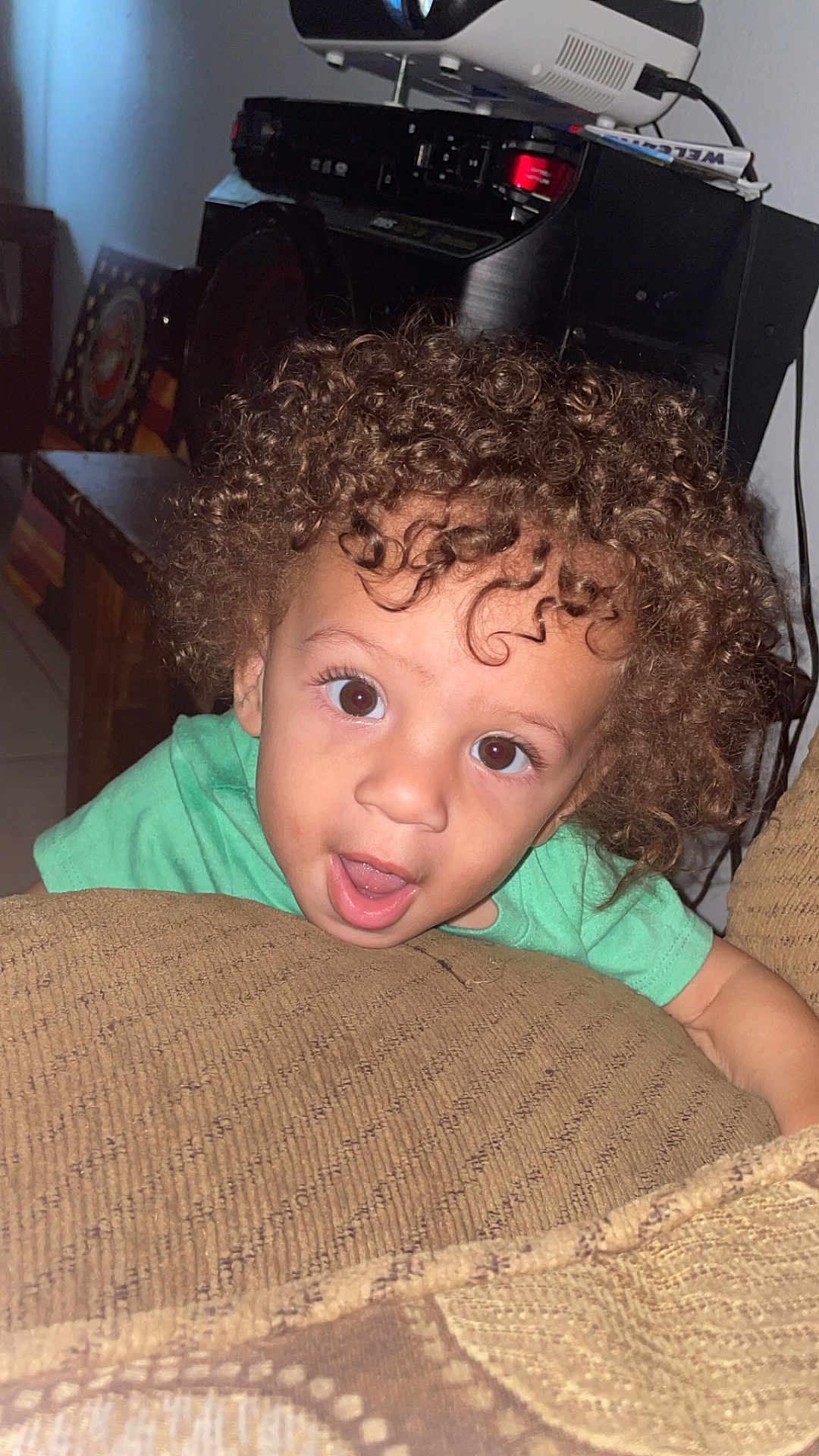 Camari joined the competition — help win amazing prizes! child, toddler, curly_hair, green_shirt, couch, indoor, face, wide_eyes, open_mouth, curious, living_room, furniture, cushion, brown, skin, person, young, expression, hair, smiling
