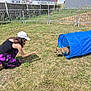 agility_tunnel, animal, blue_tunnel, casual_clothing, child, daytime, dog, fence, fun, grass, greenery, hat, leash, outdoor, pet, play, shoes, sunny, training, woman