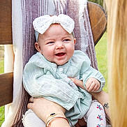 Reagan is registered to the contest to win money with this photo: baby, child, smiling, headband, bow, outdoor, greenery, blanket, wooden_chair, person, holding, tattoo, bracelet, long_sleeve, socks, happy, portrait, daylight, cute, infant