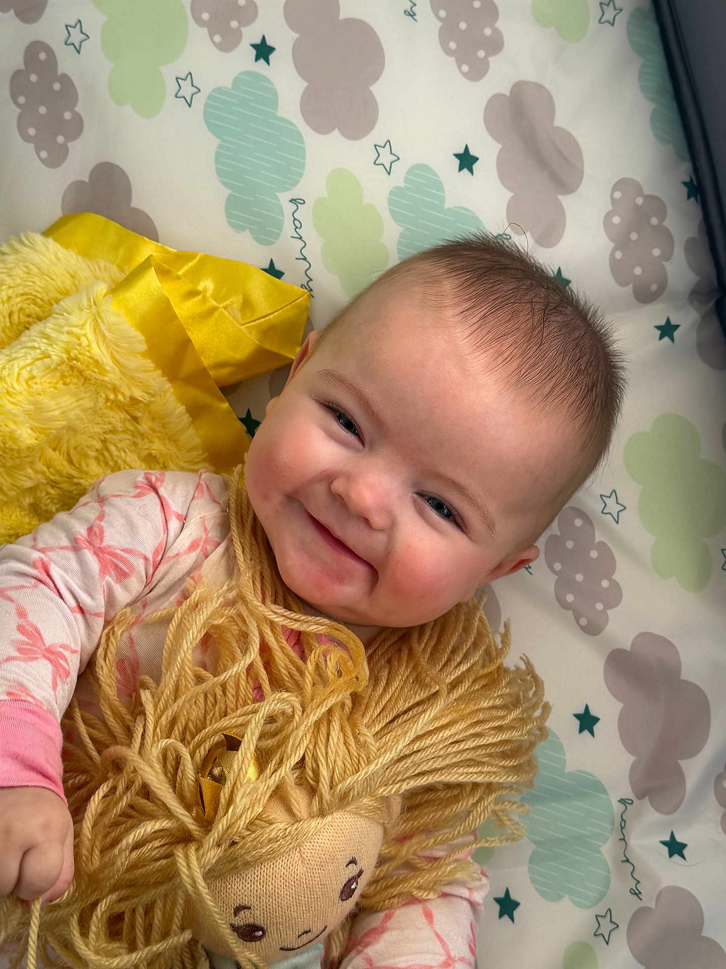 Reagan joined the competition — help win amazing prizes! baby, smiling, doll, yarn_hair, plush_toy, blanket, cloud_pattern, star_pattern, infant, child, cute, happy, soft_toy, lying_down, indoors, pink_clothing, yellow_blanket, face, person, portrait