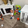 Gian is registered to the contest to win money with this photo: toddler, child, toy, basketball_hoop, ball, floor, cabinet, vase, roses, indoor, playtime, curious, kneeling, colorful, wooden_floor, white_cabinet, furniture, person, smiling, home