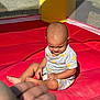 toddler, child, inflatable, play_area, red, sunlight, shadow, hand, sitting, outdoor, yellow, grey_outfit, pattern, baby, expression, summer, cute, person, playtime, young