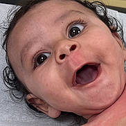 Chozen is registered to the contest to win money with this photo: baby, blanket, child, closeup, curly_hair, cute, expression, eyes, face, happy, human, indoor, infant, lying_down, mouth, open_mouth, person, portrait, skin, soft_surface