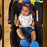 baby, car_seat, hat, bib, toys, blue_clothing, indoor, child, infant, curious, seat, person, footwear, expression, colorful, wood_floor, casual, snapshot, phone_interface, wearing