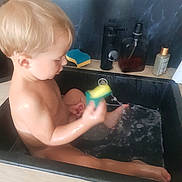 Victor a rejoint le concours — aidez-le/la à gagner de superbes lots ! toddler, child, sink, water, bath, sponge, kitchen, faucet, soap_dispenser, playing, wet_skin, bath_time, sitting, splashing, blond_hair, candid, indoor, young_child, cleaning, fun