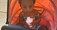 Nyuri is registered to the contest to win money with this photo: baby, child, cup, curious, curly_hair, cute, daylight, expression, food, outdoor, person, pink, playful, shade, sitting, small, stroller, summer, sunlight, toddler