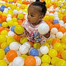 ball_pit, blue_balls, child, colorful_balls, curly_hair, cute, entertainment, fun, indoor, kid, orange_balls, pink_sweater, play_area, playful, portrait, smiling, toddler, white_ball, yellow_ball, young_child