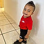 toddler, child, smiling, nike, red_shirt, black_shorts, mohawk, standing, indoor, tile_floor, white_wall, black_shoes, necklace, happy, person, casual_clothing, portrait, young_child, fashion, cute