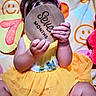 baby, child, yellow_dress, milestone, seven_months, cardboard_sign, hands, legs, headband, colorful_blanket, smiley_faces, sitting, indoors, cute, infant, clothing, person, celebration, portrait, announcement