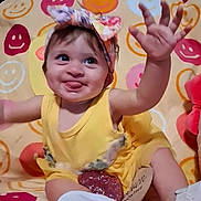 Brynlee is registered to the contest to win money with this photo: baby, child, yellow_dress, headband, tongue_out, smiling, hand, sitting, colorful, blanket, face, cute, playful, indoors, footwear, white_shoes, expression, happy, person, infant
