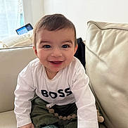 Qayim a rejoint le concours — aidez-le/la à gagner de superbes lots ! baby, child, infant, toddler, smiling, face, eyes, hair, white_shirt, text_on_shirt, couch, sofa, living_room, indoor, portrait, happy, sitting, pacifier_clip, cushions, adorable