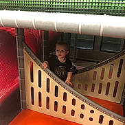 Michael is registered to the contest to win money with this photo: child, toddler, playground, indoor_playground, play_structure, mesh_netting, orange_floor, safety_padding, panel, slide, metal_pole, candid, curious, shirt, nike_logo, hand_on_panel, seat, window, soft_play, portrait
