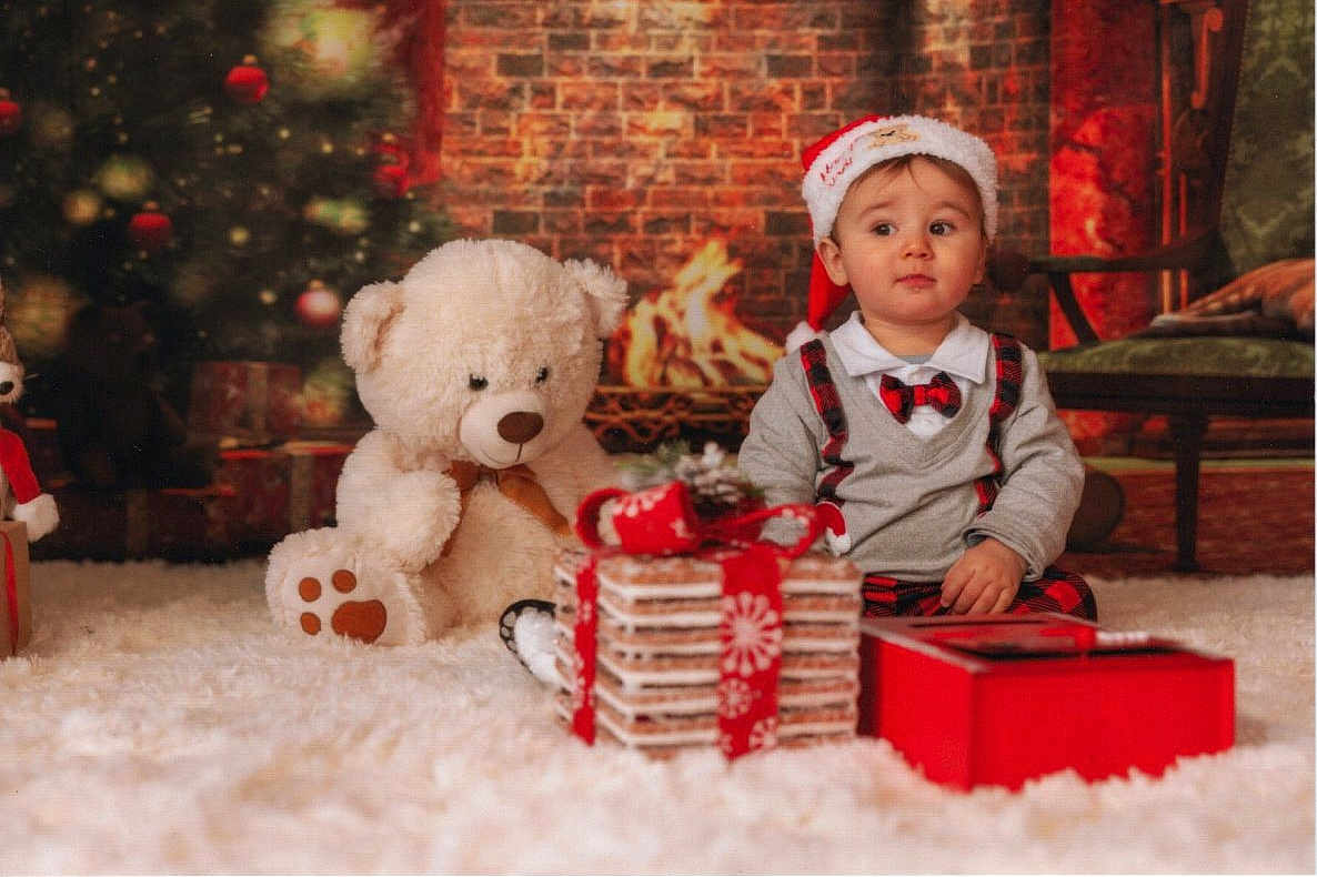 Julian participe au concours pour gagner de l'argent avec cette photo : baby, carpet, child, christmas_tree, cozy, cute, decorations, festive, fireplace, gift, holiday, indoors, plaid, portrait, present, red, santa_hat, sitting, teddy_bear, white