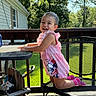 toddler, child, smile, pink_clothing, outdoor, deck, metal_chair, table, sunlight, greenery, house, happy, playful, summer, daylight, railing, shadows, socks, young_child, balancing