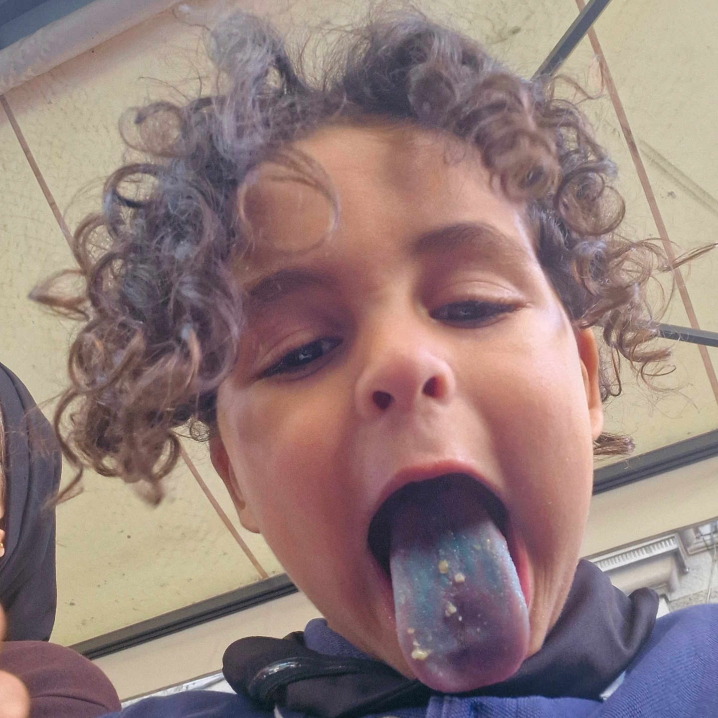 Emanallah a rejoint le concours — aidez-le/la à gagner de superbes lots ! blue_tongue, buttoned_shirt, casual_clothing, child, close_up, crumbs, curly_hair, daylight, expression, face, fun, head, mischievous, outdoor, person, playful, portrait, roof, tongue_out, young