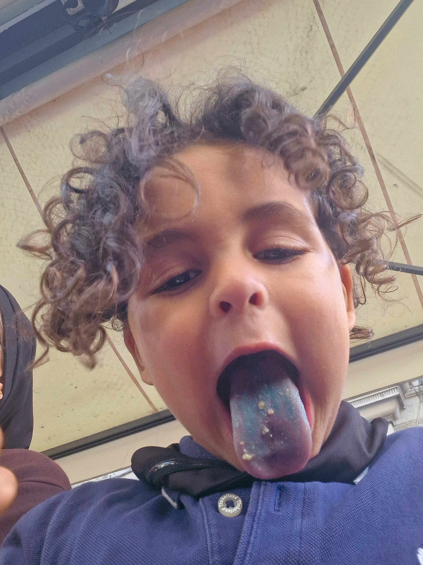 Emanallah a rejoint le concours — aidez-le/la à gagner de superbes lots ! child, curly_hair, blue_tongue, crumbs, playful, face, tongue_out, casual_clothing, buttoned_shirt, outdoor, roof, mischievous, close_up, young, person, portrait, fun, expression, head, daylight