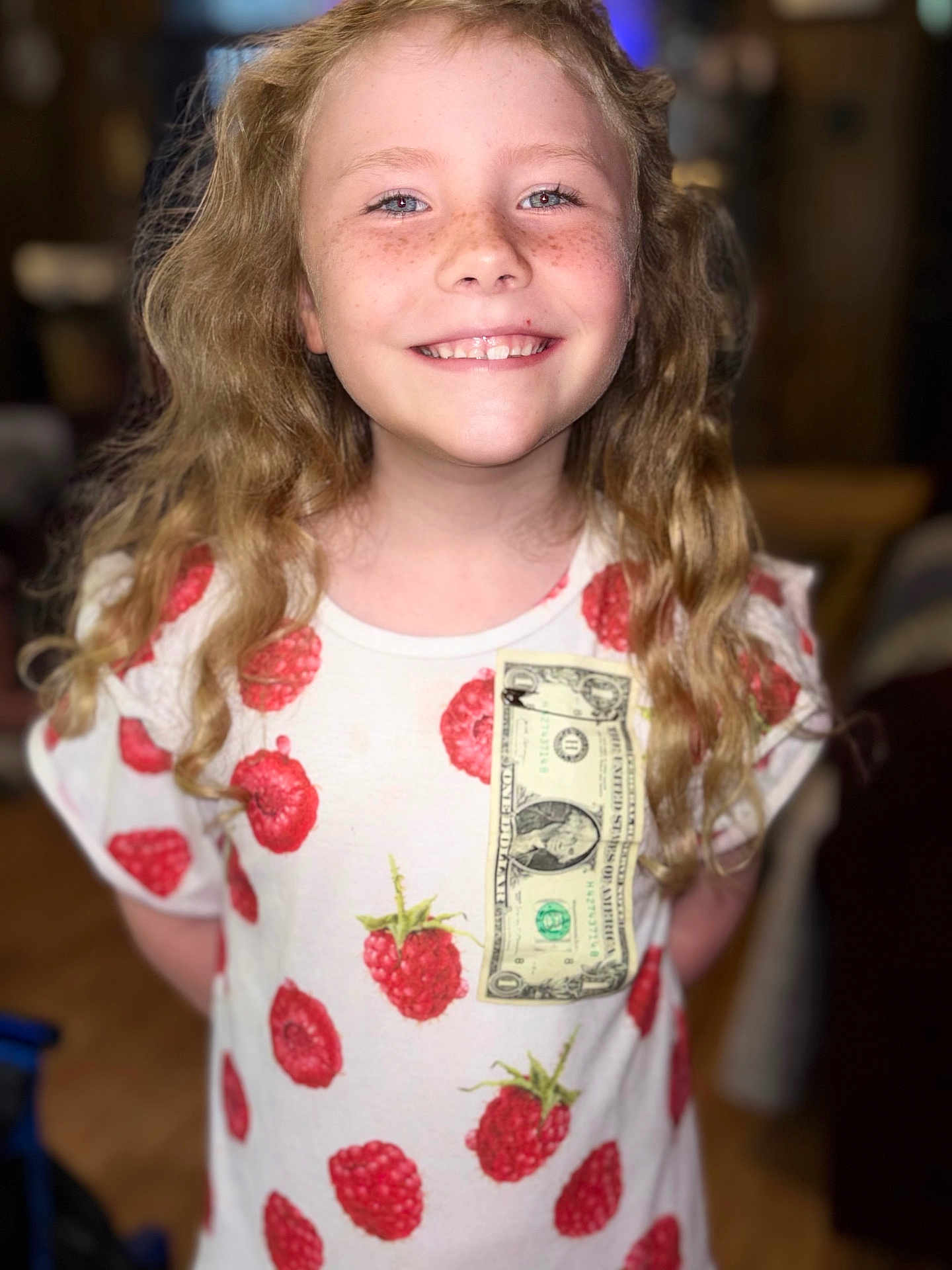Willow is registered to the contest to win money with this photo: child, smiling, freckles, blonde_hair, curly_hair, tshirt, raspberry_pattern, dollar_bill, indoor, portrait, happy, person, face, casual_clothing, young, cute, standing, childhood, fashion, closeup