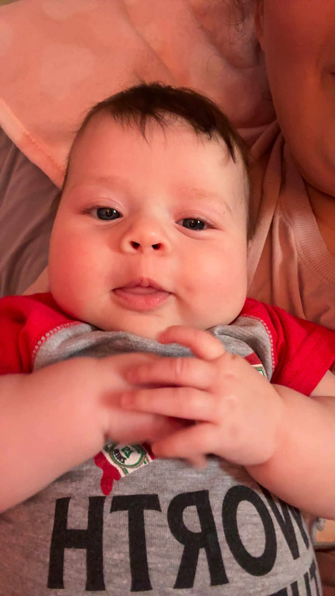 Scarlett is registered to the contest to win money with this photo: baby, infant, child, face, hands, red_shirt, gray_shirt, text_on_clothing, blanket, soft_texture, person, portrait, closeup, cute, smile, indoor, skin, head, young_child, expression