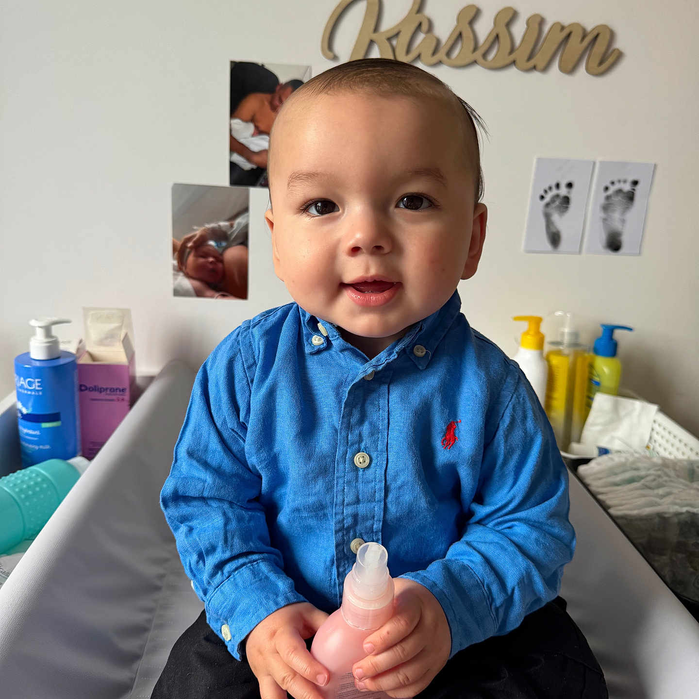 Kassim participe au concours pour gagner de l'argent avec cette photo : baby, child, boy, blue_shirt, black_pants, bottle, sitting, nursery, photographs, footprints, name_sign, indoor, smiling, face, hands, clothing, person, cute, portrait, young