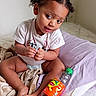 toddler, child, bed, blanket, pillow, lipton, bottle, hair_clip, curly_hair, white_wall, indoor, diaper, tshirt, sitting, cross_legged, soft_texture, curious_expression, natural_light, cozy, young_child