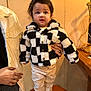 toddler, child, jacket, checkered_pattern, white_shoes, standing, indoor, person, hand, support, pants, brown_hair, cute, warm_lighting, chair, casual, background, smiling, fashion, portrait