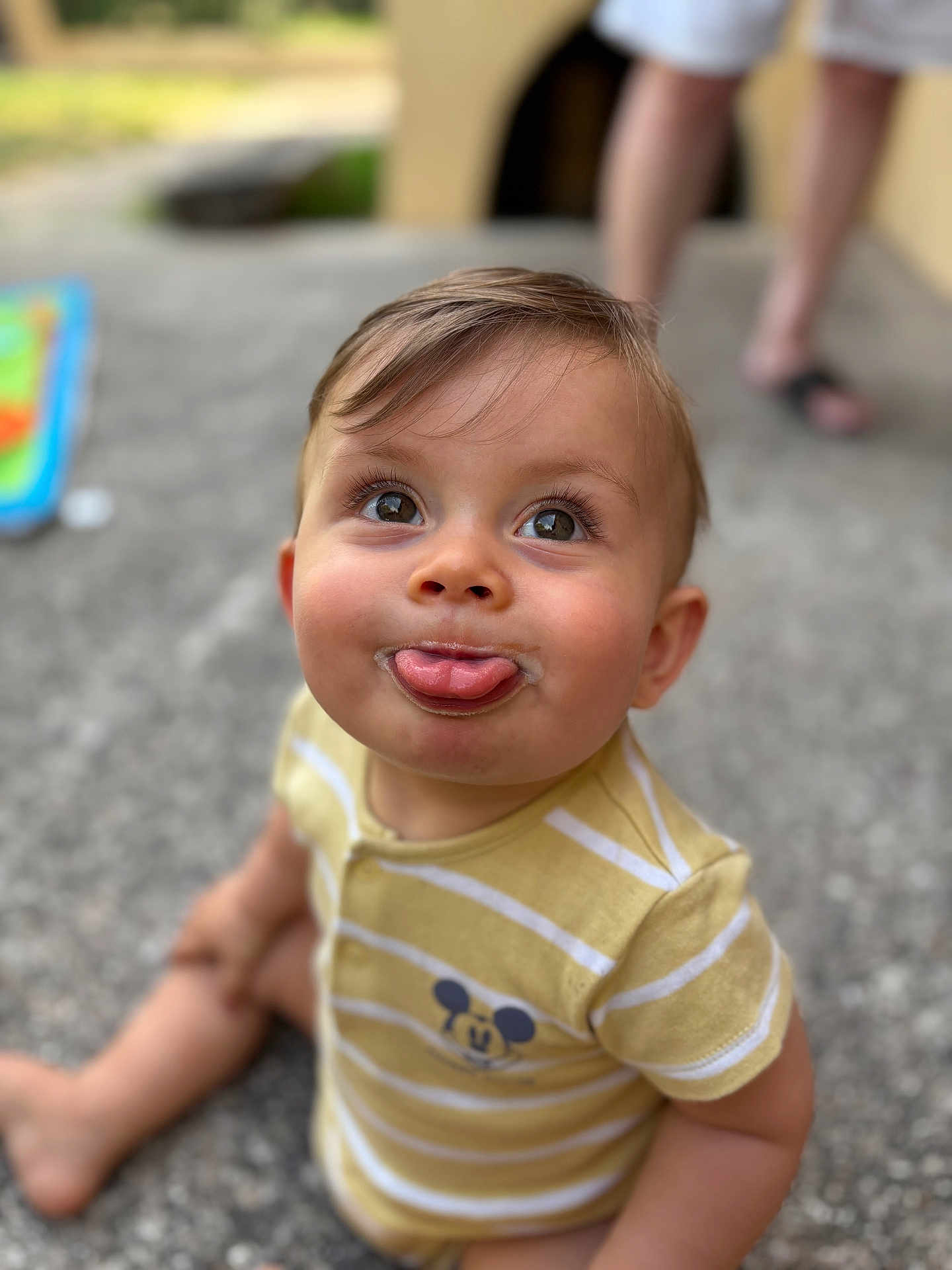 Ayden participe au concours pour gagner de l'argent avec cette photo : toddler, child, baby, tongue_out, yellow_clothing, striped_shirt, mickey_mouse, sitting, concrete_floor, outdoor, playful, cute, face, expression, person, legs, blurred_background, summer, shorts, barefoot