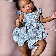 Reigna Sky is registered to the contest to win money with this photo: baby, infant, child, dress, patterned_clothing, wooden_buttons, lying_down, pink_background, curly_hair, smiling, cute, skin, hand, foot, portrait, indoors, soft_texture, fabric, closeup, person